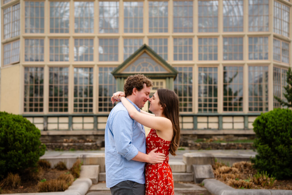 Couple embracing and smiling in front of the Rawlings Conservatory greenhouse in Baltimore, MD during an engagement photo session
